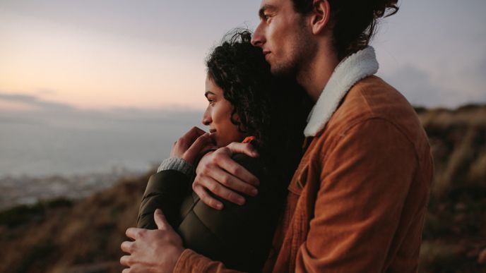 Romantic young couple standing in mountain together and looking at view. Young man embracing his girlfriend and looking away.