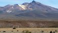 Bílá branka v pouštním písku na pozadí žluto-cihlovo-červené siluety vulkanických úbočí Barevných hor (Montanas Coloradas) je jak z palety futuristického malíře.