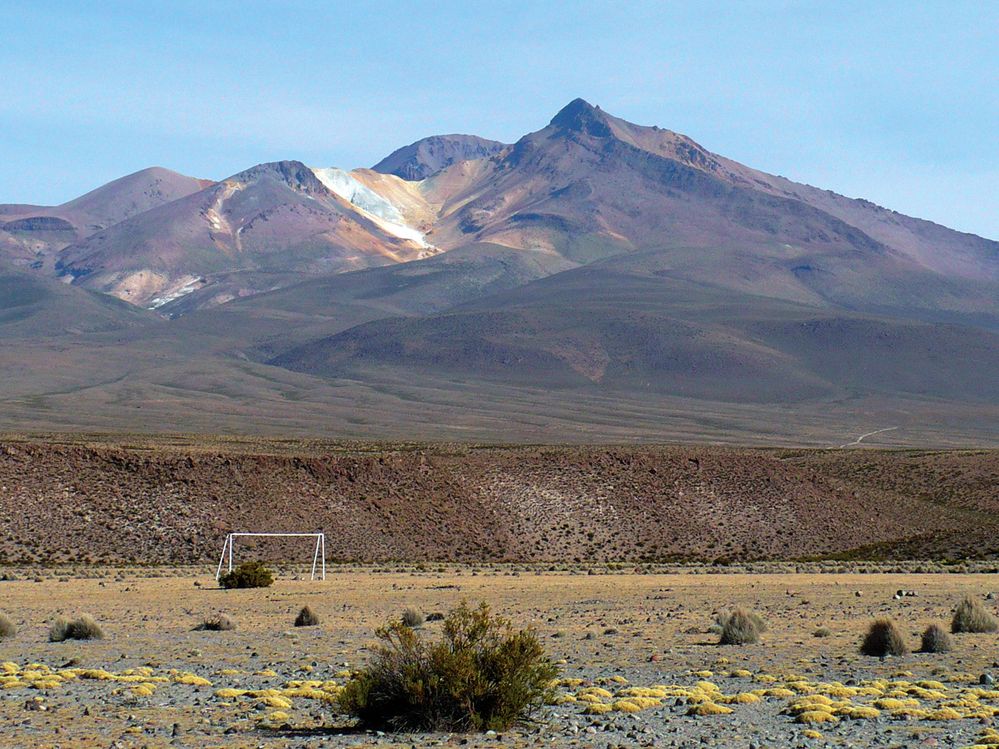 Bílá branka v pouštním písku na pozadí žluto-cihlovo-červené siluety vulkanických úbočí Barevných hor (Montanas Coloradas) je jak z palety futuristického malíře.