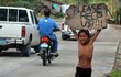 Chlapec stojí u silnice a nad hlavou drží ceduli s nápisem »Prosím, pomozte nám!!!!«.