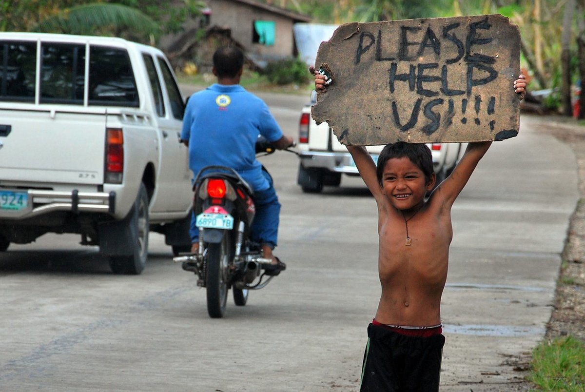 Chlapec stojí u silnice a nad hlavou drží ceduli s nápisem »Prosím, pomozte nám!!!!«.