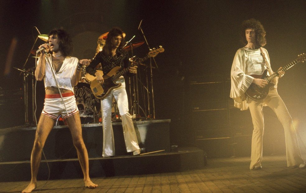 Kapela Queen. Freddie Mercury u mikrofonu, po jeho levici vlasáč Brian May. Jeho odhadované jmění je 210 milionů dolarů.