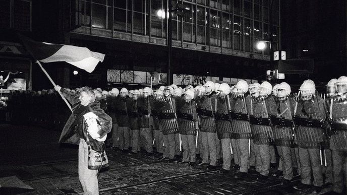 Počátek. Žena se státní vlajkou 17. listopadu 1989demonstruje na Národní třídě před policejním kordonem.Policie vzápětí proti studentům brutálně zasáhla