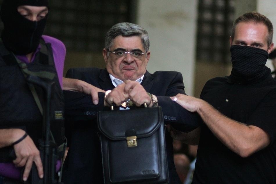 Řecký politik Nikos Michaloliakos se netváří zrovna dvakrát šťastně. Vedou ho do policejní cely.