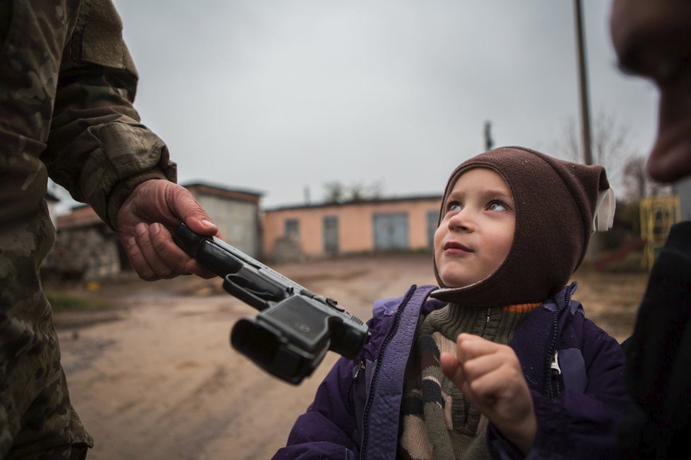 Muž podává zbraň dítěti, aby si zkusilo ten pocit, držet zbraň. „War isn’t over yet – women in war and others“ (Válka ještě neskončila – ženy a ti další ve válce)