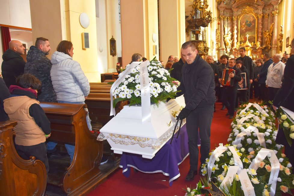 Bílou rakev se zastřelenou ženou odvážejí pracovníci pohřební služby kolem květy z kostela ven.