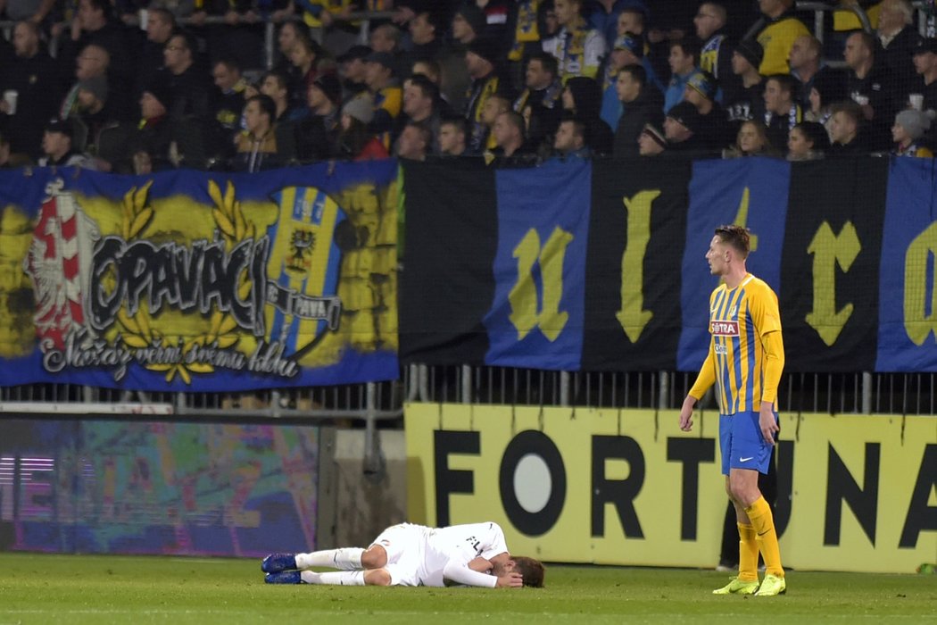 Plzeňský záložník Tomáš Hořava leží v bolestech na zemi, vpravo přihlíží opavský Dominik Simerský