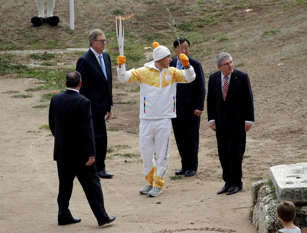 Biatlonista Apostolos Aggelis vybíhá na první úsek olympijské štafety, zatímco ho sleduje šéf MOV Thomas Bach a další funkcionáři