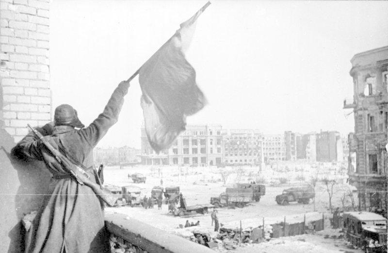 Sovětský voják mává rudým praporem v troskách města.