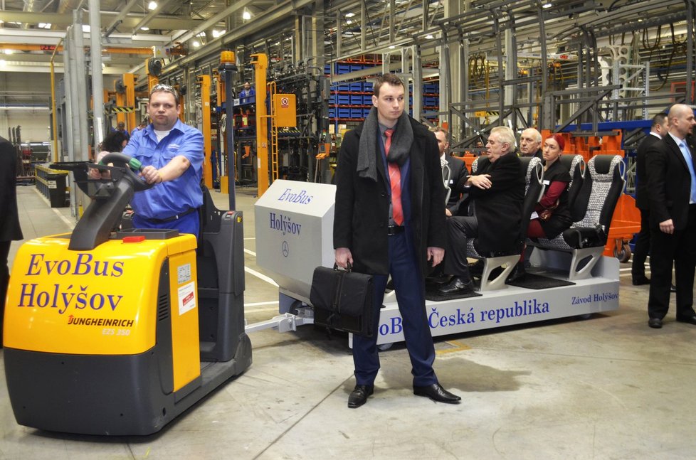Březen 2015, Holýšov na Domažlicku: Muž s taškou chodí vedle prezidenta, který jede »ještěrkou« při návštěvě v továrně na výrobu autobusů.